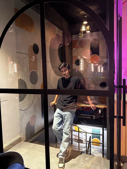 A young man in a black t-shirt and light blue jeans poses leaning against a railing inside what appears to be a restaurant or cafe with decorative glass panels and warm pendant lighting.