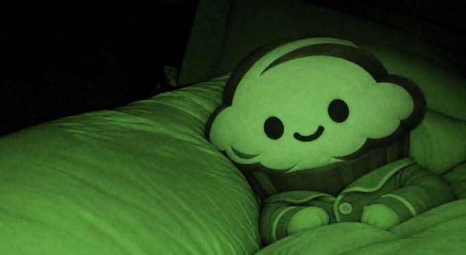 A glowing plush toy shaped like a smiling cupcake character rests on a pillow in a dark room, bathed in green nightlight. The cute, meme-worthy image pairs with the post's playful misspelling of "start" as "$tart," suggesting the cozy mascot symbolizes a welcoming, approachable beginning for the next-generation DEX on Binance Smart Chain.