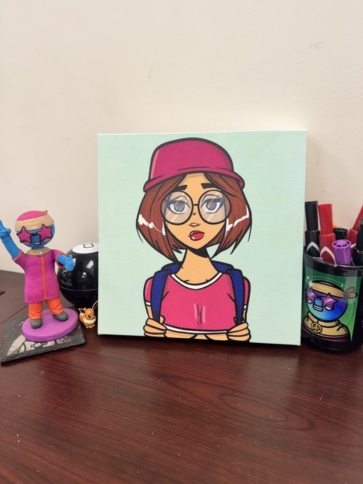 The image features a wooden desk with various items placed on top of it. There is an art piece displayed prominently on the desk, which appears to be a painting or drawing of a girl wearing glasses and a hat. The artwork is accompanied by a small figurine of a girl dressed in pink and orange clothing. In addition to these objects, there are several pens scattered around the desk, as well as a cup placed nearby. A post on social media has been shared with this image, which seems to be about Ana @theinkedminkNFT's artwork or a similar piece of art.