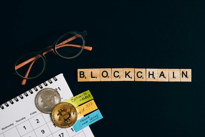 The image features a calendar with the word "Blockchain" written on it, surrounded by various objects related to cryptocurrency and digital currency. There are three coins placed around the calendar, one of which has the number "21" on it. A pair of glasses is also present in the scene, adding an element of personalization or style to the overall composition. The image seems to be promoting a message about privacy and blockchain technology, as more institutions and financial products incorporate privacy into their digital currency offerings.