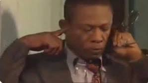 A still of a man in a suit and tie with his fingers in his ears, appearing to block out sound or noise.