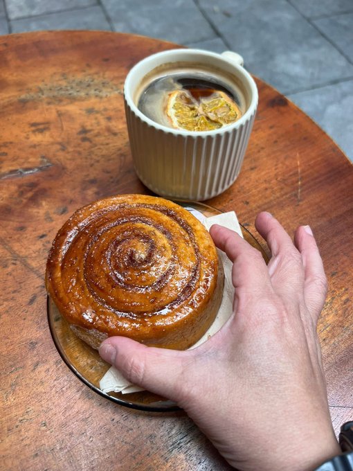 A hand reaches for a freshly baked cinnamon roll beside a cup of coffee with an orange slice, set on a round wooden café table. The scene captures the cozy Sunday morning ritual of enjoying a pastry and coffee before settling in to watch Bitcoin.
