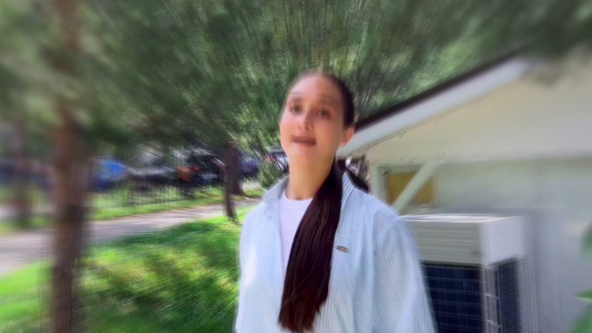 The image shows a woman with long brown hair wearing a white shirt standing outside of a house or building. The photo has been edited to create an artistic effect that makes her appear as if she's floating, giving the impression that she's looking up at something in the distance.