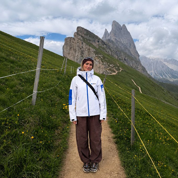 A woman wearing a white "MEXC" jacket stands on a mountain trail in the Dolomites, with the iconic jagged peaks rising behind her—fittingly showcasing how the brand's merch performs at new heights in the great outdoors.