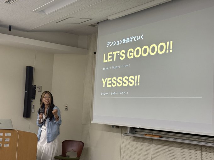 A woman stands at a podium giving a presentation, with a projected slide behind her displaying the text 'テンションをあげていく' and 'LET'S GOOOO!!' and 'YESSS!!' with Japanese translation hints.