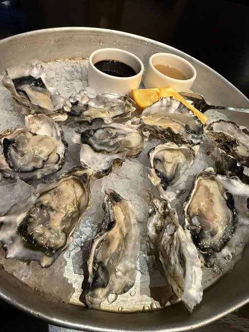 An overhead photo of a metal bowl filled with crushed ice holding a dozen raw oysters on the half shell, accompanied by two small cups of dipping sauces and lemon wedges.