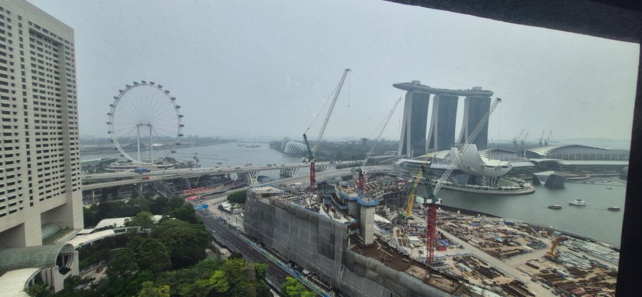A view from a high-rise window in Singapore showing the Marina Bay area, including the Singapore Flyer ferris wheel, the Marina Bay Sands hotel, the ArtScience Museum, and ongoing construction work with cranes in the foreground.