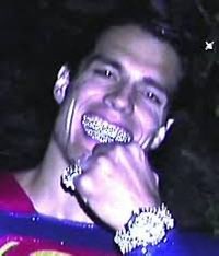 A smiling young man in a red shirt with diamond-encrusted grills and jewelry rests his hand on his chin.