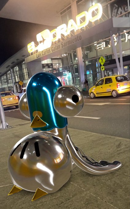 A nighttime photo of a polished chrome penguin sculpture with a teal head at El Dorado airport, with the illuminated 'ELDORADO Luis Carlos Galán Sarmiento' sign and a yellow taxi visible in the background.
