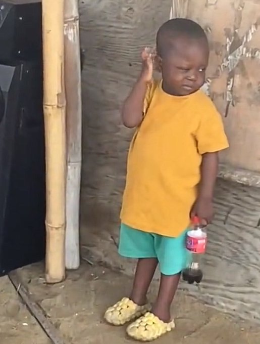 A young child wearing a yellow t-shirt, teal shorts, and yellow crocs, holding a bottle of pink soda while touching their ear and looking slightly downward.