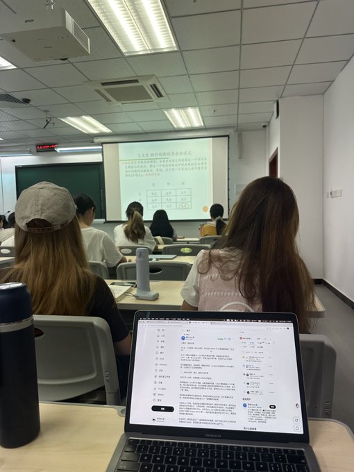 A classroom scene from the back of the room showing students seated at desks facing a projected presentation screen, with a MacBook in the foreground displaying a social media feed.