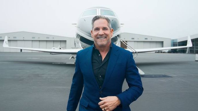 A smiling man in a blue blazer and black shirt standing in front of a private jet on an airport tarmac.
