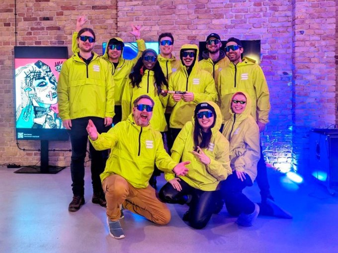 The image features a group of people posing for a photo with their arms raised in front of them, wearing yellow jackets or hoodies. The group consists of at least nine individuals, all dressed similarly in yellow clothing. They are standing next to each other against a brick wall backdrop and a large screen displaying an image of a woman's face. The text "fun fact this was the very first Build Station in existence" is also present in the image.