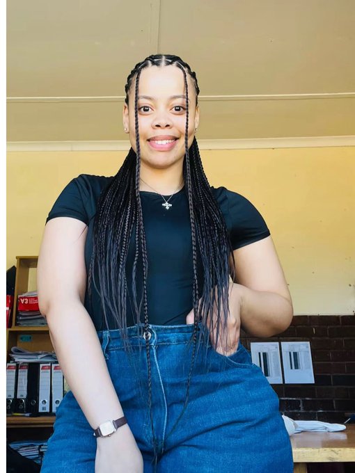 A smiling young woman with long braided hair, wearing a black short-sleeve top, blue jeans, and a silver cross necklace, poses for a portrait in what appears to be an office or classroom setting.