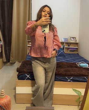 The image features a woman taking a selfie with her cell phone while standing next to a bed. She is wearing a red top and gray pants, which are laid out on the bed. The room has a brown color scheme, including a brown comforter on the bed. There is also a potted plant in the background, adding some greenery to space.