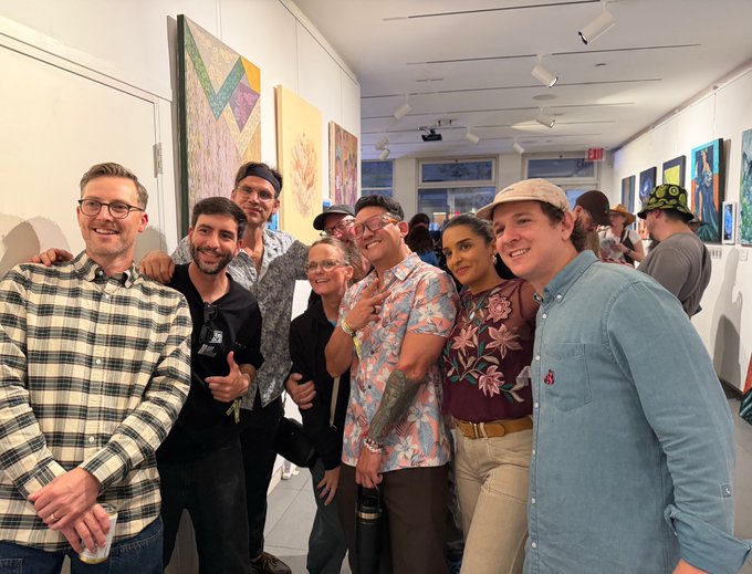 A group of seven people smiling and posing together at what appears to be an art gallery opening, with colorful paintings displayed on the walls behind them.