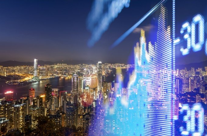 A nighttime cityscape featuring Hong Kong's skyline with skyscrapers illuminated along the harbor, overlaid with glowing blue digital numbers and financial charts.