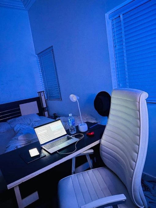 A blue-lit bedroom workspace features a white ergonomic chair at a black desk with an open laptop, a desk lamp, and a bed visible in the background.