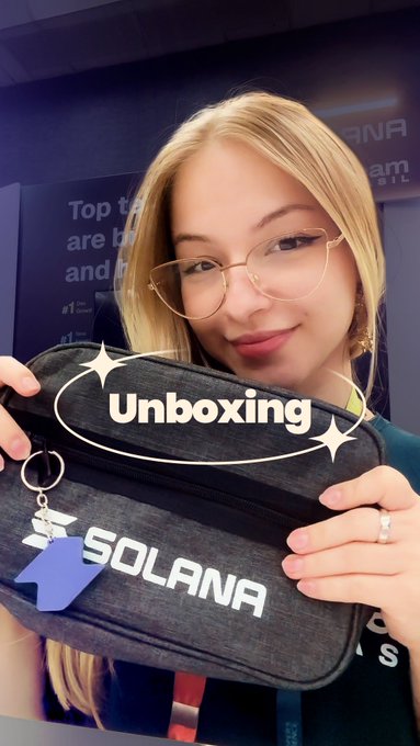 A young woman with blonde hair and glasses smiles while holding up a black denim pouch featuring the "SOLANA" logo and a keychain. An "Unboxing" graphic overlay appears across the image, aligning with the post about unveiling the kit she developed for SuperteamBR x Solana members.