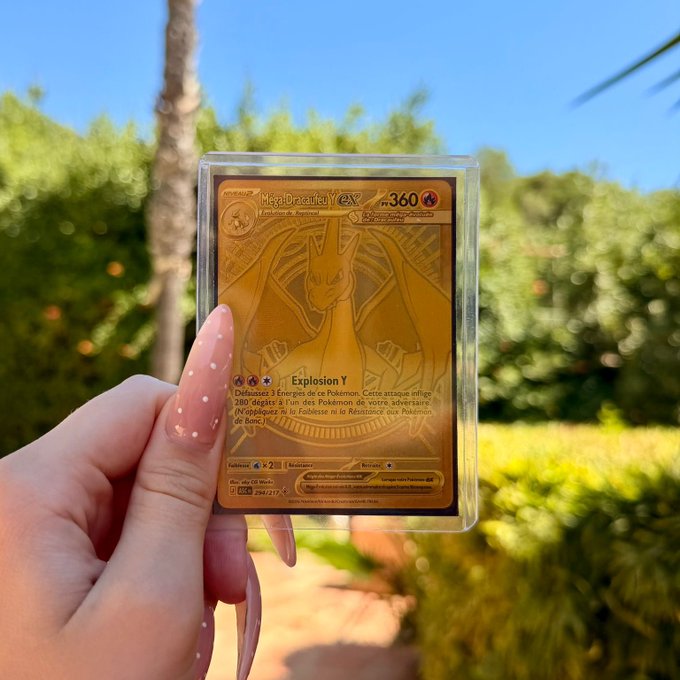 A hand with pink polka-dot nails proudly displays a gold "Méga-Dracaufeu Y GX" Pokémon card (HP 360) in a protective sleeve against a sunny garden backdrop—fitting for a "HOLY GRAIL!!" moment as this rare French gold metal card is one of the most coveted prizes in the Pokémon TCG.
