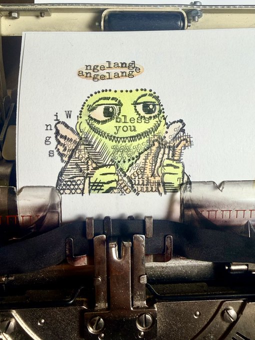 A typewriter types out a whimsical illustration of a green frog character with the words 'angelange' and 'wings bless you' visible on the paper.
