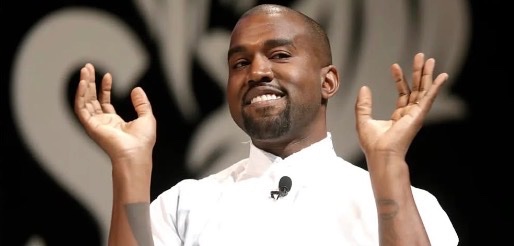A photograph of Kanye West wearing a white shirt and a headset microphone, smiling with his hands raised in the air against a dark background.