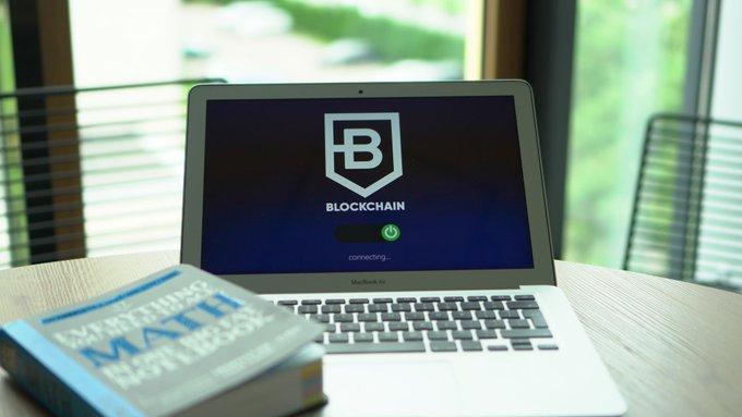 A laptop on a desk displaying a blue 'BLOCKCHAIN' logo screen with 'connecting...' text, alongside a math book titled 'MATH' in the foreground.