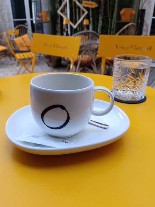 A white coffee cup with a black 'O' logo on a yellow table at a Veuve Clicquot café, with a glass of water beside it.