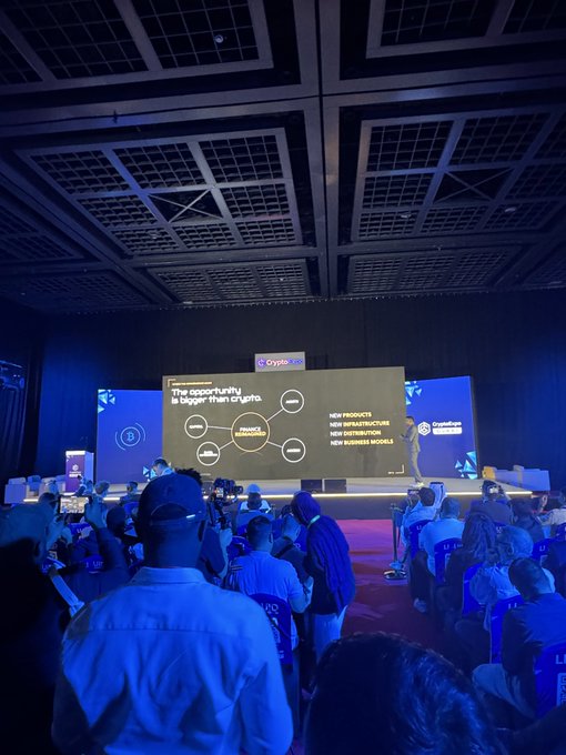 A conference scene with an audience watching a presentation slide about crypto, displaying the text 'The opportunity is bigger than crypto' with branches for new products, new blockchains, new distribution, and new business models.