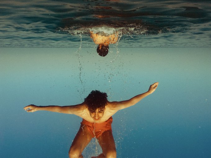 An underwater photograph capturing a man in orange swim shorts swimming toward the camera, with another person visible at the water's surface above him.