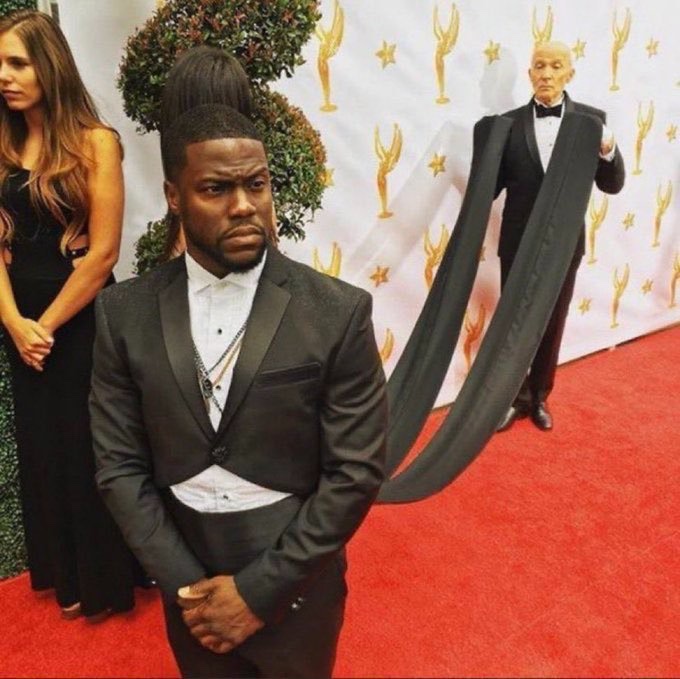 Kevin Hart stands on a red carpet looking unamused, while in the background a person in a Trump mask with dramatically elongated arms walks by at what appears to be an Emmy Awards event.