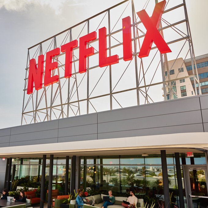 The iconic red "NETFLIX" logo towers above a modern glass-fronted building where people can be seen working on laptops in a sunlit lounge area. The image ties to the news of Netflix's upcoming limited series "The Altruists," which dramatizes the FTX scandal involving Sam Bankman-Fried and Caroline Ellison.