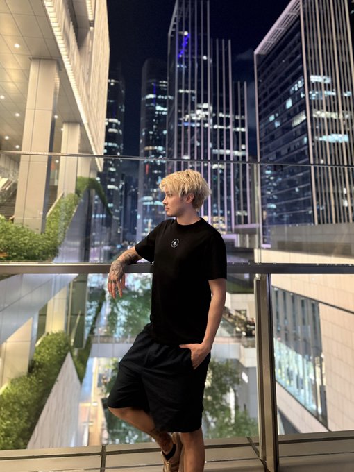 A young man with blonde hair wearing a black t-shirt and shorts leans against a glass railing on a balcony overlooking a modern cityscape with illuminated skyscrapers at night.