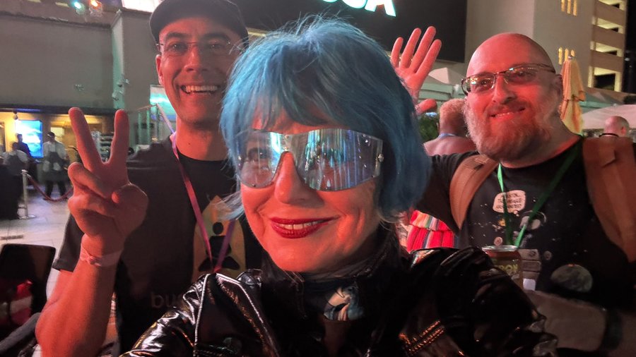 Three people posing at what appears to be a convention; the central figure has blue hair and futuristic visor sunglasses making a peace sign.