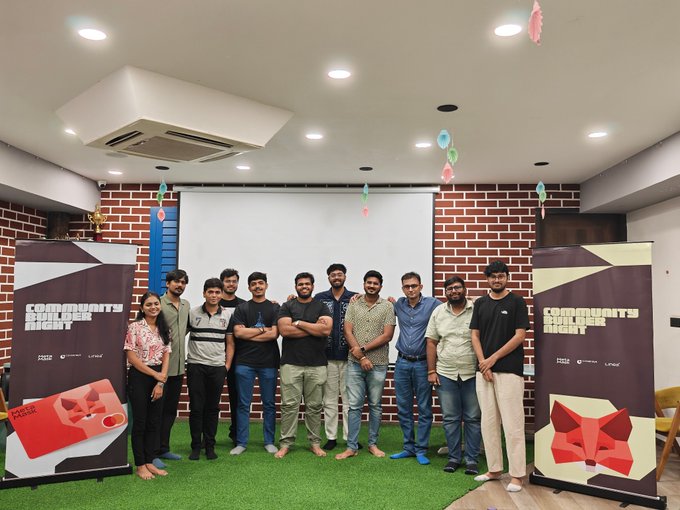 A group of about ten people posing together indoors at what appears to be a 'Community Build Night' event, standing between two promotional banners for the event.