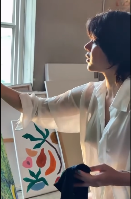 An AI-generated or stylized image of a young woman with short dark hair wearing a sheer white blouse, standing in a sunlit room near a window with a colorful abstract painting visible behind her.