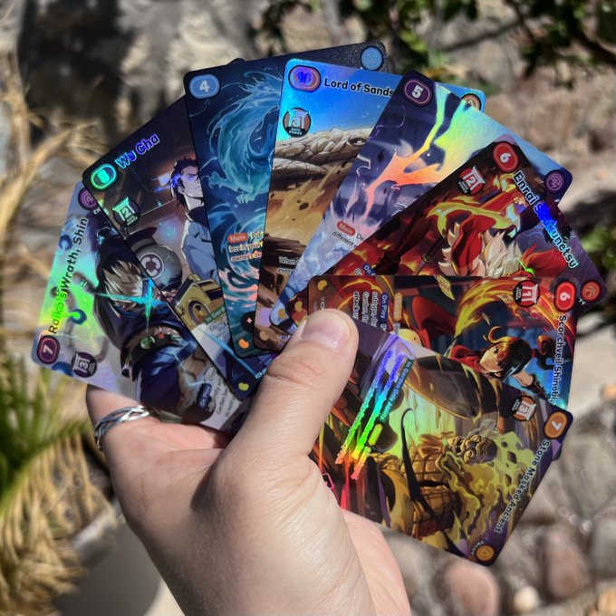 A hand holds a fan of holographic trading cards outdoors, with visible titles including 'Relic Wraith, Shin', 'Wu Glu', 'Lord of Sands', and 'Eternal Shogun' (Sakazaki).