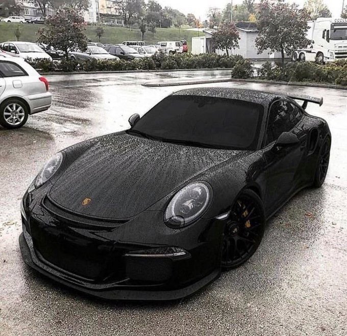 A matte black Porsche 911 sports car with a large rear wing and custom wide body kit parked in a wet lot on a rainy day.