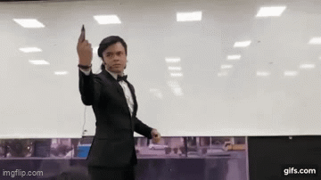 The image shows a man in a black tuxedo and bowtie standing in an office setting, holding up a phone triumphantly with a confident smirk, evoking a "V for Victory" pose. The "Day 1 of posting source verified facts until MOASS🚀" caption suggests the man is celebrating sharing the first verified fact in a daily series, with his victorious pose symbolizing conviction and excitement for the upcoming MOASS (Mother of All Short Squeezes) event.