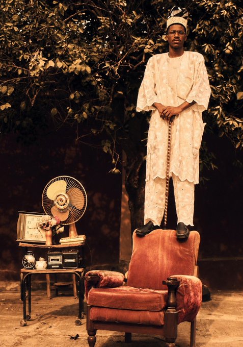 A sepia-toned photograph shows a man in white traditional clothing and cap standing on top of a red armchair, with a vintage fan, clock, and radio beside him.
