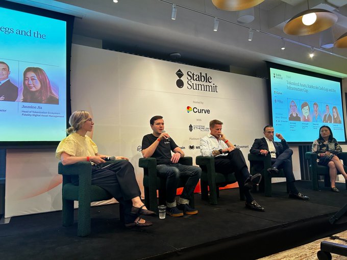The image shows a group of five people sitting around a table, engaged in a discussion or presentation. They appear to be at an event where they are discussing the concept of digital currency and its potential impact on major tradfi institutions. The panelists include two women and three men, all seated comfortably on chairs arranged around the table.

The room has a large screen behind them that displays information related to their discussion or presentation. There is also a water bottle placed near one of the participants' seats.