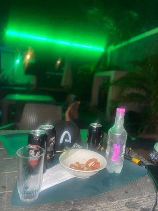A dimly lit indoor scene with green LED lighting, featuring a table with soda cans, a glass, a bowl of tomato slices, and a pink-capped water bottle in the foreground.