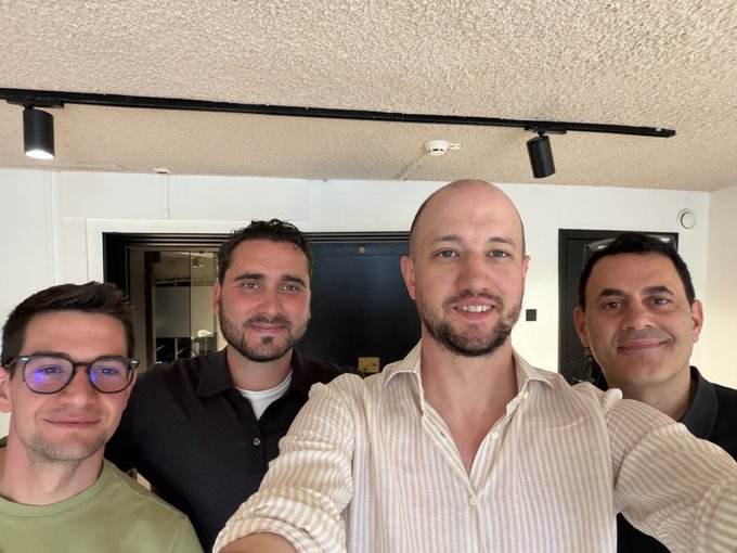 The image features four men standing next to each other, posing for a photo with smiles on their faces. They are all wearing glasses and appear to be friends or acquaintances. The background of the image shows a white wall, which contrasts nicely with the black ceiling in the room where they are standing. There is also an elevator visible in the scene, suggesting that this gathering might have taken place in an office building or similar setting.