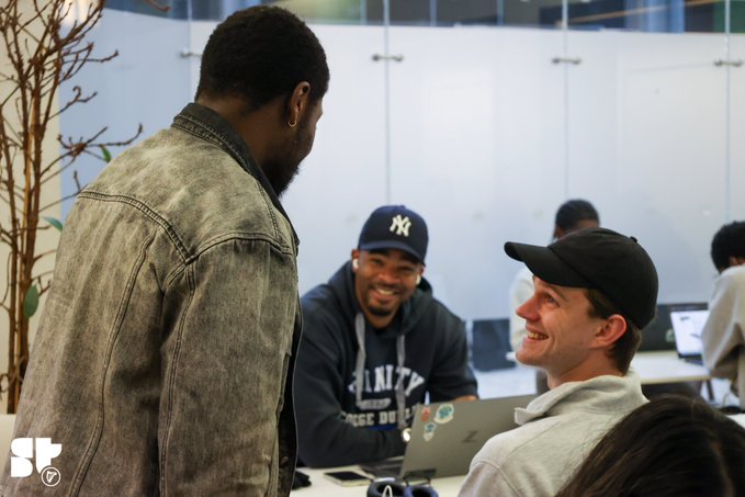 Three young men converse and smile in an office or classroom setting, one wearing a 'Brooklyn College District' hoodie and NY Yankees cap, with an 'ST' watermark in the corner.