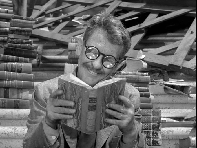 A black-and-white image of a man with round glasses smiling widely while reading an open book, surrounded by stacks of old books.