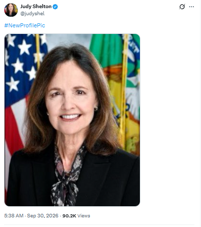 A social media post by Judy Shelton (@judyshel) featuring her professional headshot with an American flag and green official flag in the background, tagged #NewProfilePic.
