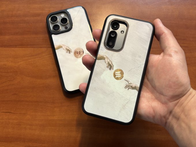 A person holds a smartphone featuring a case printed with the 'Creation of Adam' artwork, where a Bitcoin coin replaces the space between the two hands.