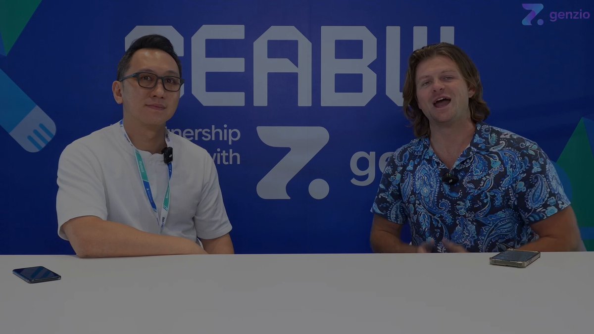 The image shows two men sitting behind a table with their cell phones placed on top. They are positioned at a booth during an event, possibly a conference or panel discussion. One of them has a name tag on his shirt that reads "David ThaDegen". The other man is wearing a blue and white patterned shirt. The backdrop features a large blue banner with the text "SEABWofficial" written in white letters.