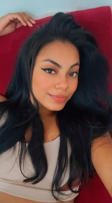 A selfie of a young woman with long black hair and winged eyeliner lying on a red cushion, wearing a light pink/beige top.