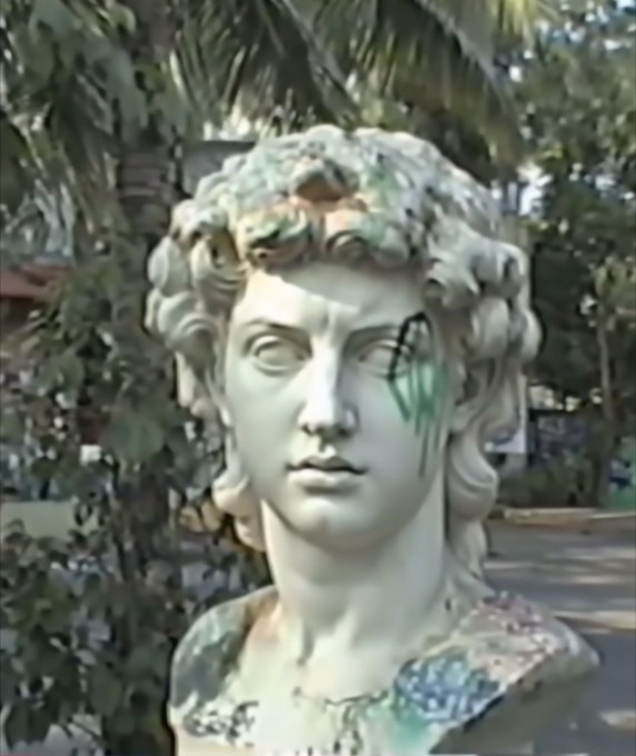 A white marble statue bust with curly hair has been vandalized with green spray paint dripping down the right side of its face, set against a backdrop of palm trees.